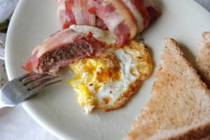 Fork cutting into bacon wrapped around ham and sausage on plate with egg and toast.